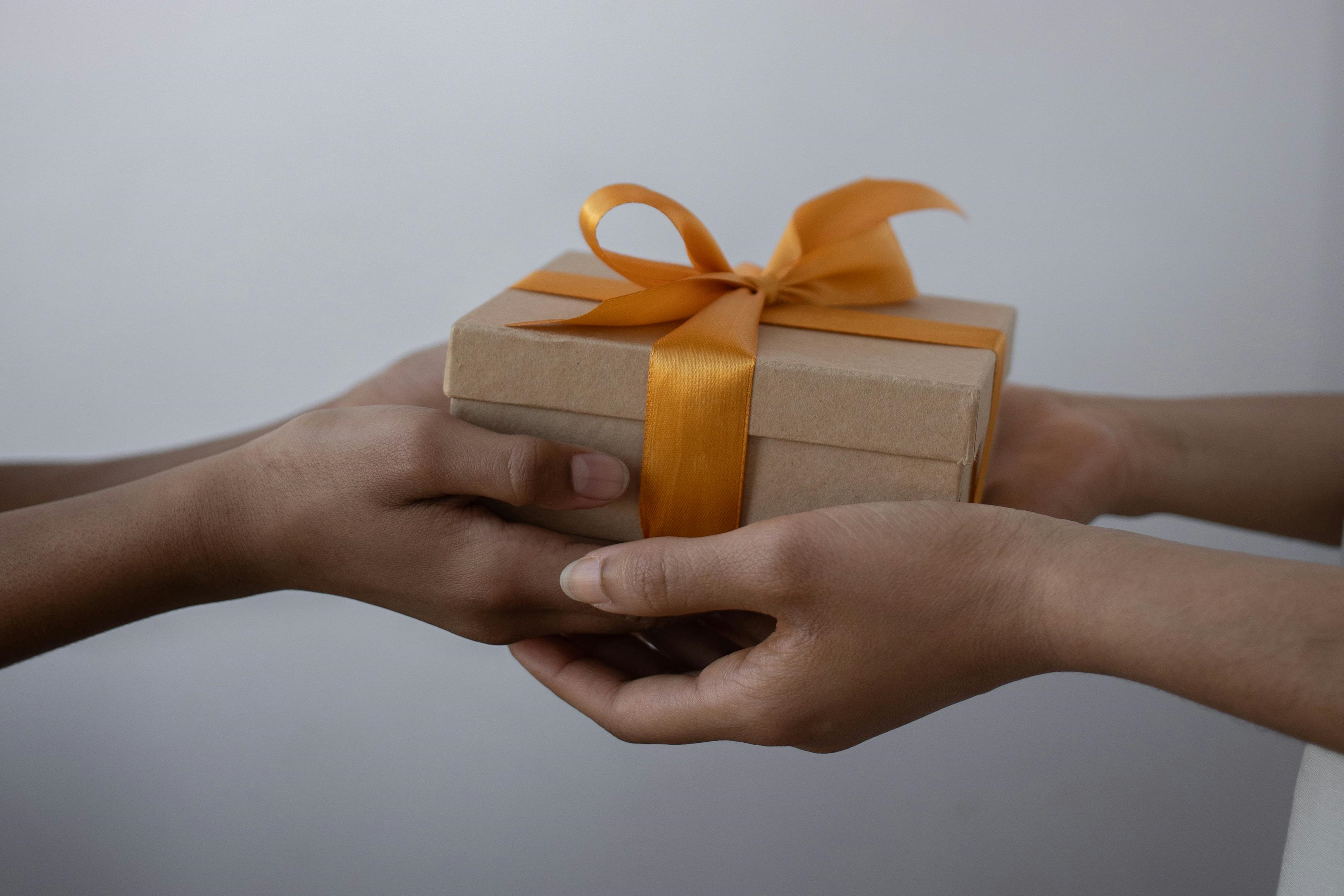 Two hands exchanging a gift: the receiving hand is below and the giving hand is above, holding a gift box